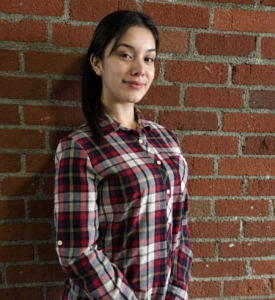 Portrait of a young woman with long dark hair wearing a red, navy, and white plaid button-up shirt, standing against a red brick wall with a slight smile. Warm natural lighting highlights her face, creating a friendly and approachable professional portrait.